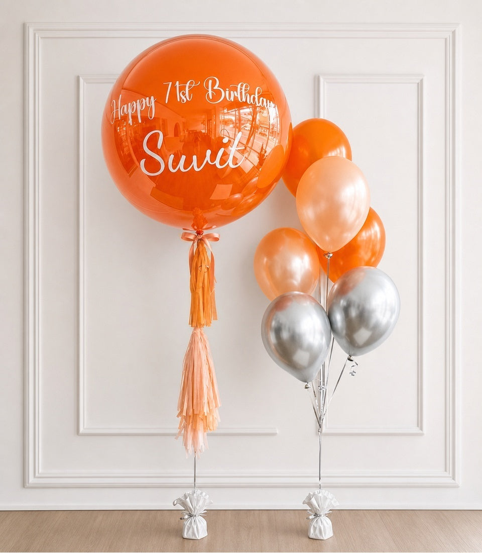 Orange and silver foil balloons with tassel decoration and "Happy 1st Birthday Swidi" text on large bubble balloon.