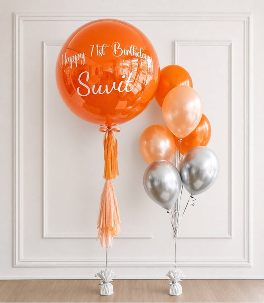 Orange and silver foil balloons with tassel decoration and "Happy 1st Birthday Swidi" text on large bubble balloon.