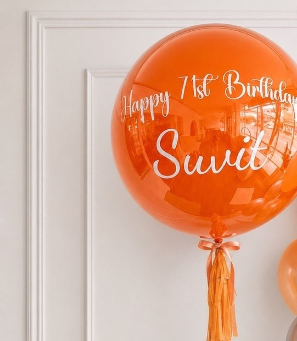 Orange spherical foil balloon with white text reading "Happy 1st Birthday Swati" and orange tassel attached to bottom against white wall.