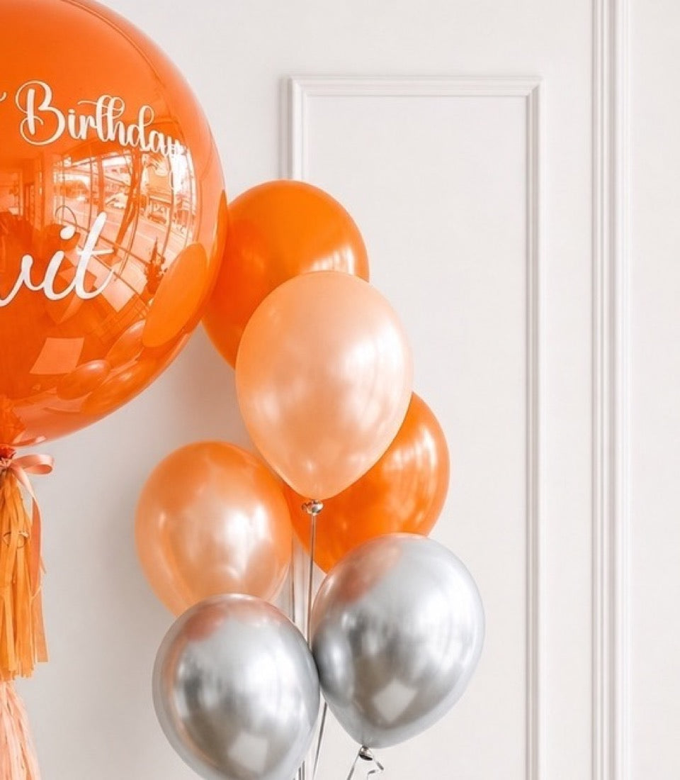 Orange and silver metallic balloons clustered together with a large "Birthday" bubble balloon, against a white wall with framed door.