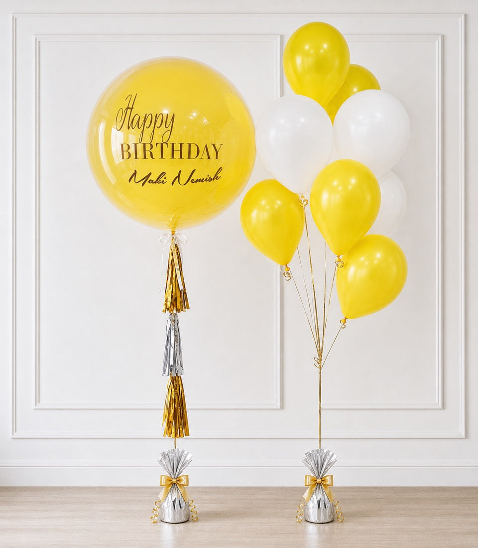 Yellow and white balloons with gold tassels and bows against white paneled wall. Large yellow bubble balloon reads "Happy Birthday Maki Nomish."