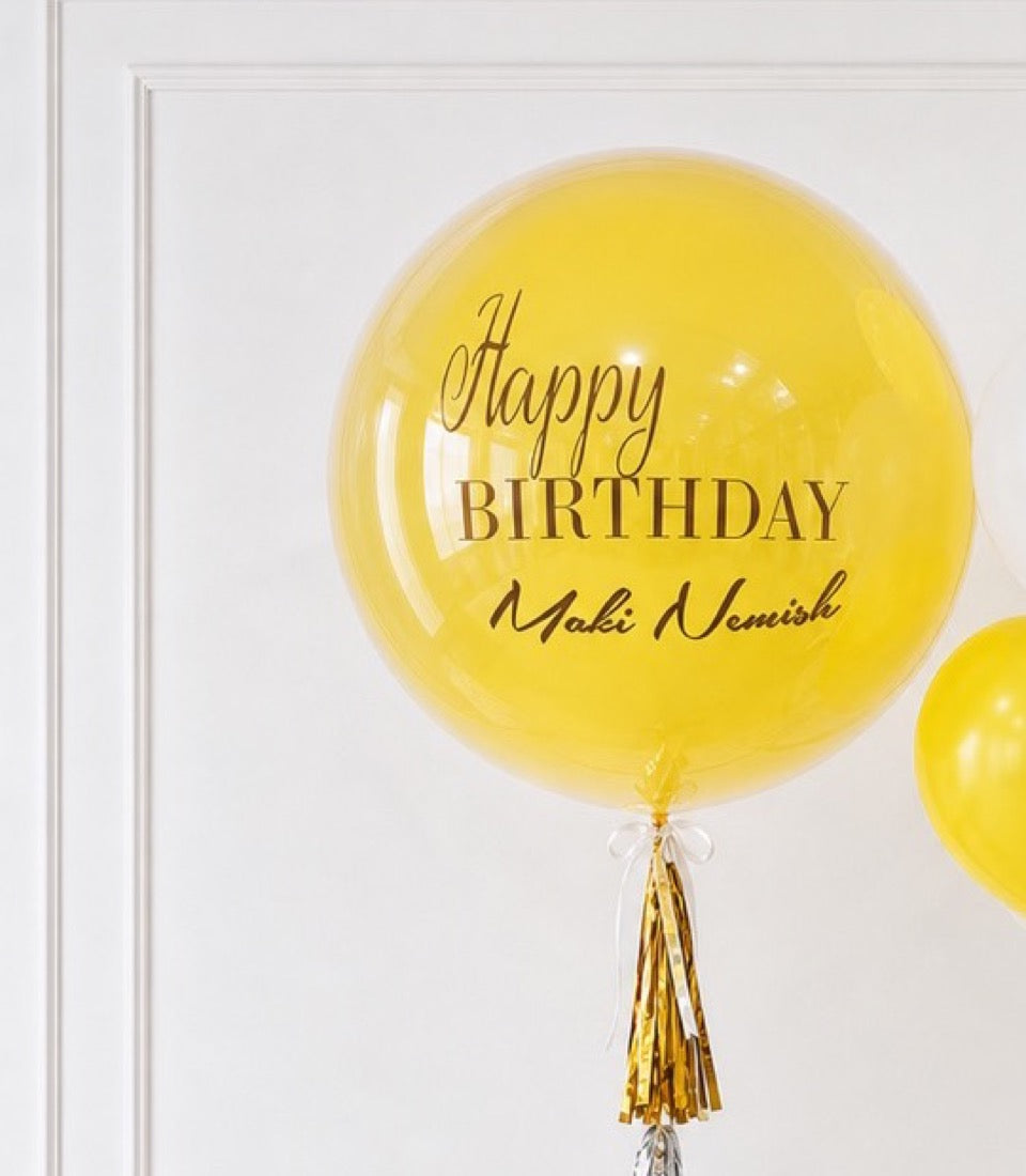 Yellow personalized bubble balloon with "Happy Birthday" and "Maki Nemish" text, gold tassel, against white wall.