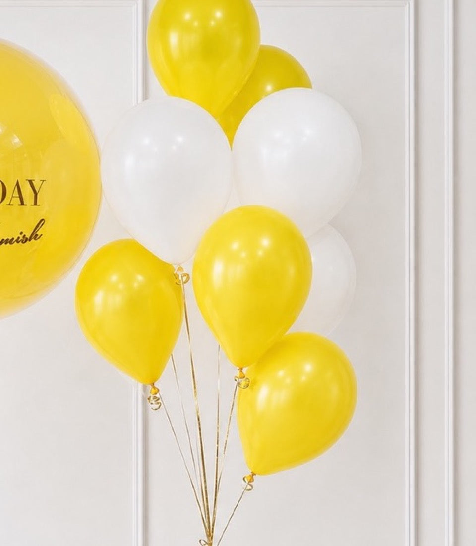 Yellow and white balloons with gold ribbons clustered together against a white paneled wall, featuring a large yellow balloon with text on the left.
