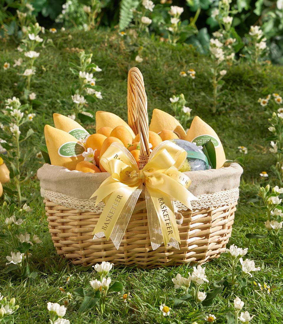 Wicker gift basket filled with ripe yellow mangoes, orange fruit, and gourmet treats, lined with beige fabric and white lace