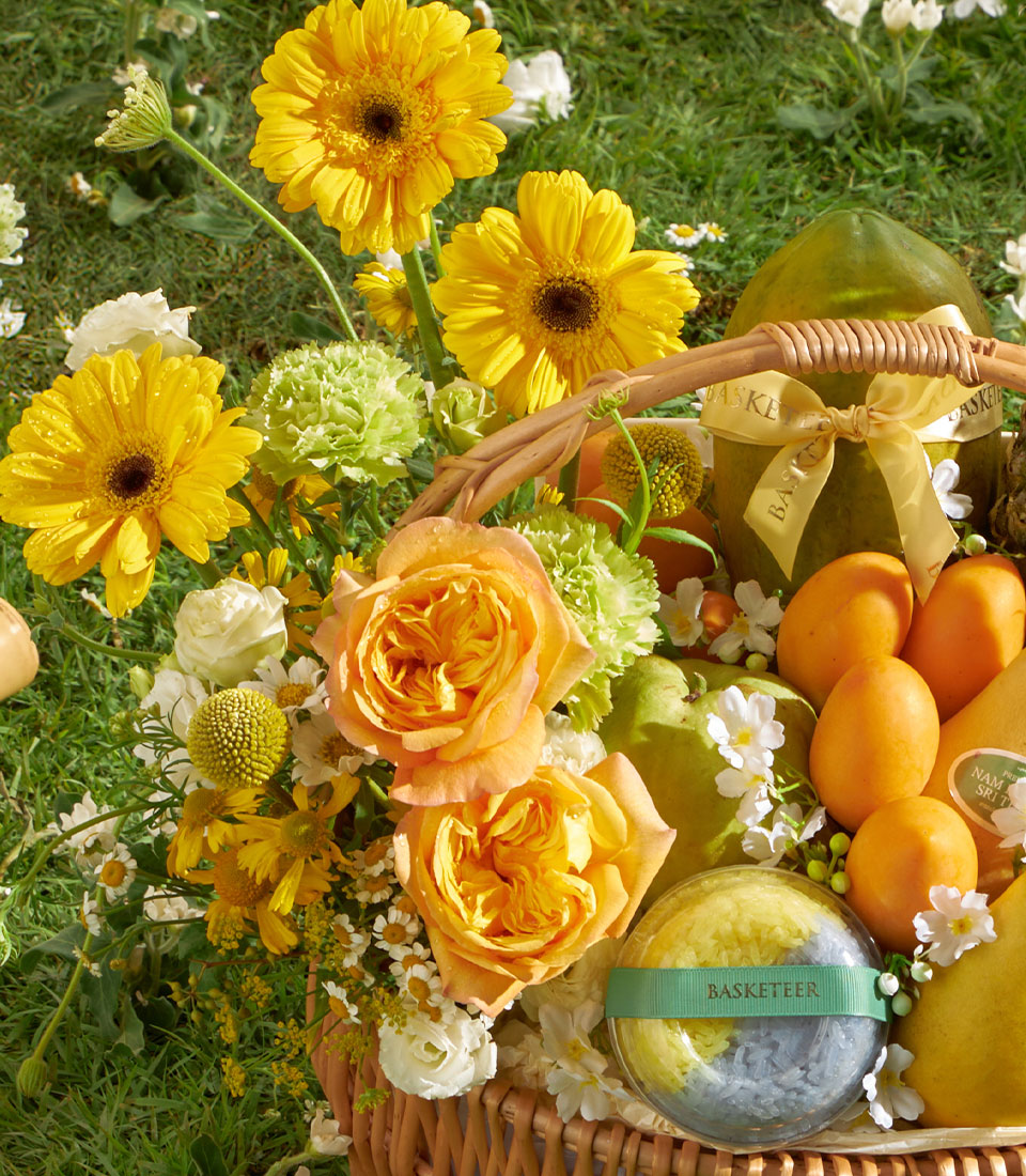 Sunlit Mango Garden gift basket with yellow gerbera daisies, orange roses, fresh mangoes, papaya, and pears on green grass