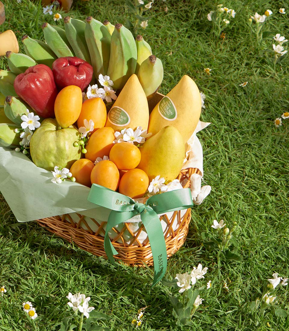Woven wicker basket overflowing with fresh Thai mangoes, bananas, guavas, and citrus on green tissue and white flowers