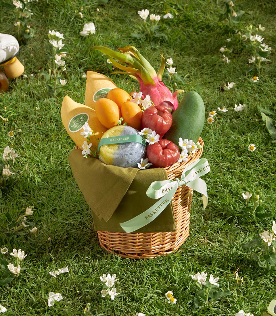Compact wicker gift basket with fresh tropical fruits, yellow mangoes, dragon fruit, and rose apples on green lawn