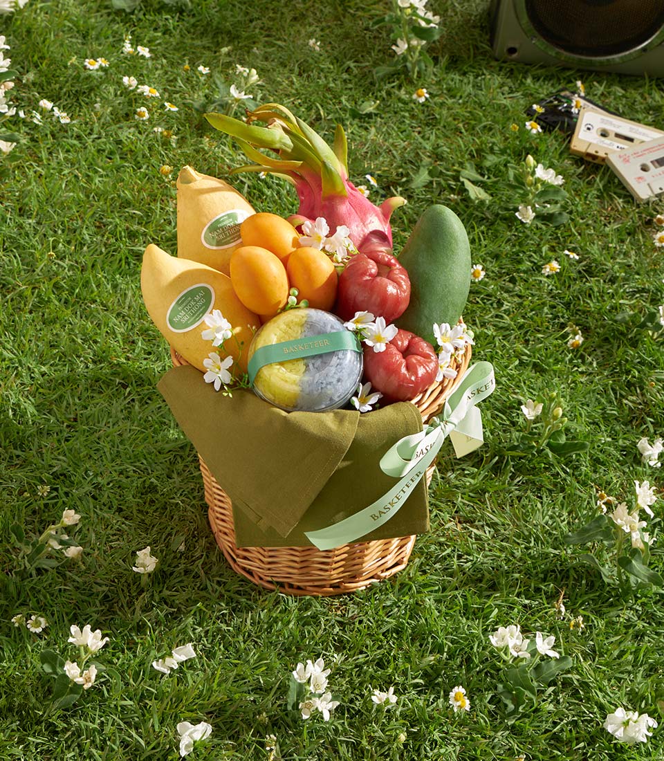 Wicker gift basket with fresh mangoes, dragon fruit, apricots, and rose apples on green lawn with white flowers