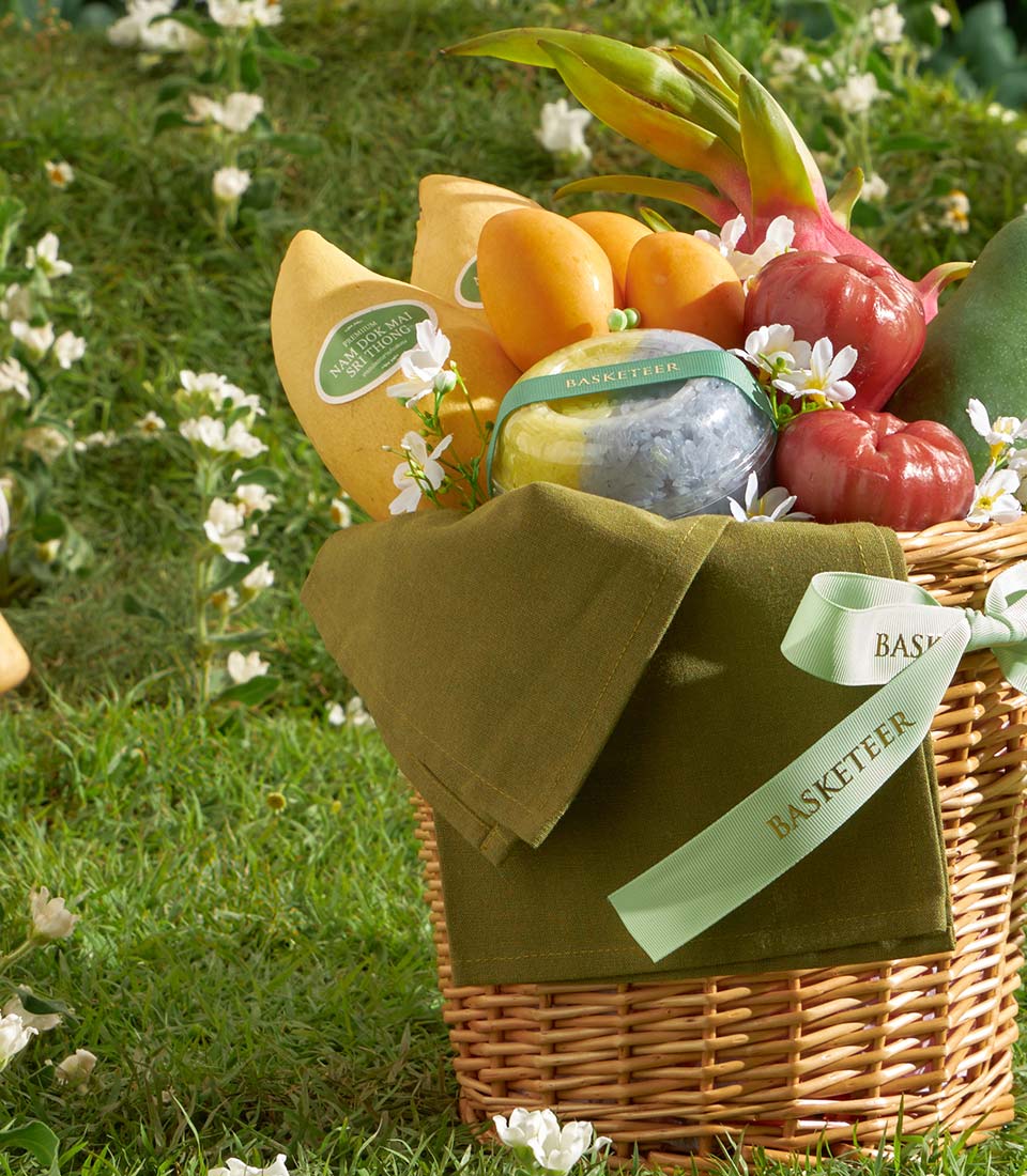 Woven wicker gift basket with fresh mangoes, layered dessert, pomegranates, and green ribbon on grass with white flowers