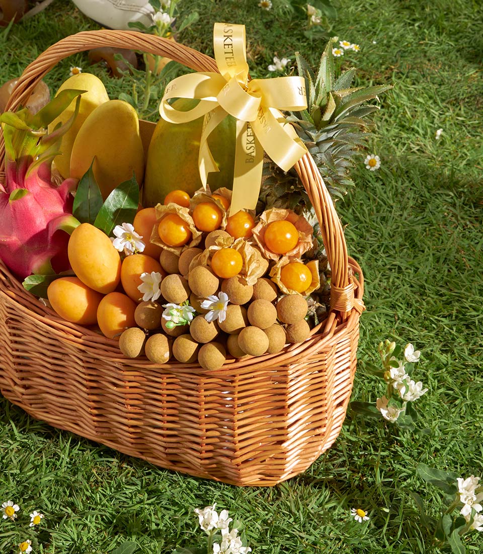 Woven wicker basket overflowing with fresh tropical fruits including mangoes, dragon fruit, papaya, and longans, tied with