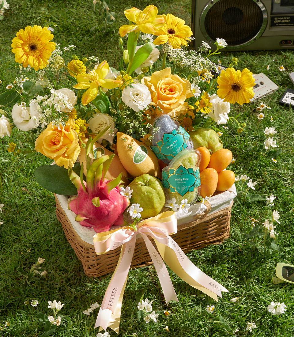 Woven wicker basket overflowing with fresh tropical mangoes, dragon fruit, guavas, and a vibrant bouquet of yellow gerbera