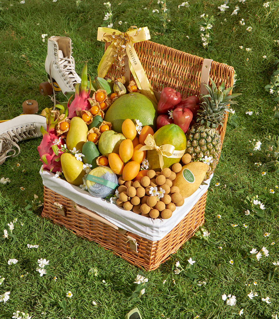 Wicker picnic basket overflowing with fresh mangoes, pomelo, longan, dragon fruit, and seasonal tropical fruits on green