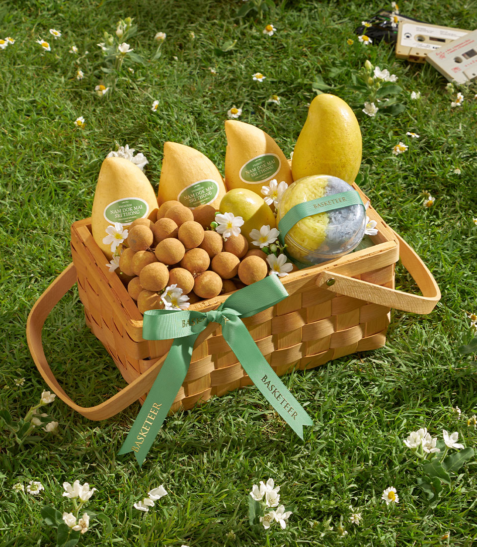 Woven picnic basket with fresh yellow mangoes, brown longans, and sticky rice dessert on green lawn with white flowers