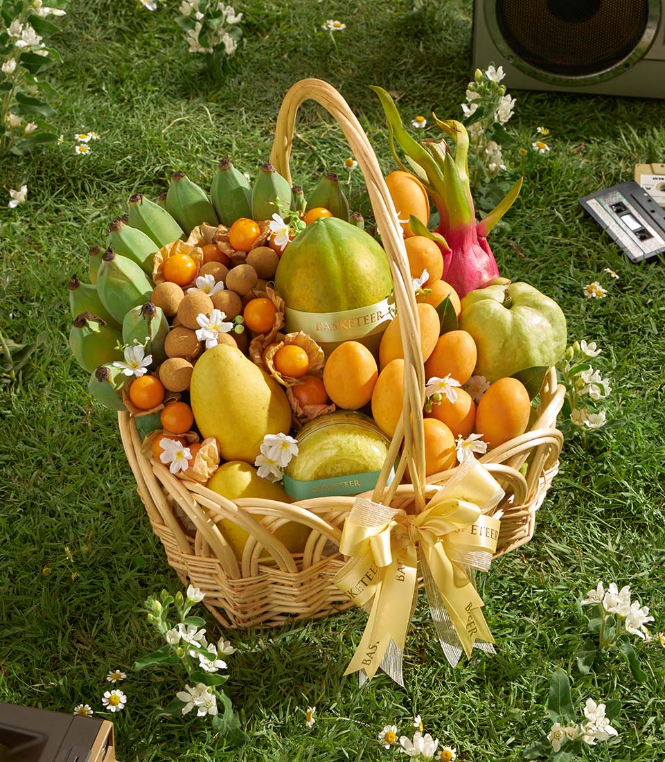 Woven wicker basket overflowing with fresh Thai tropical fruits including mango, papaya, longan, and guava on green grass