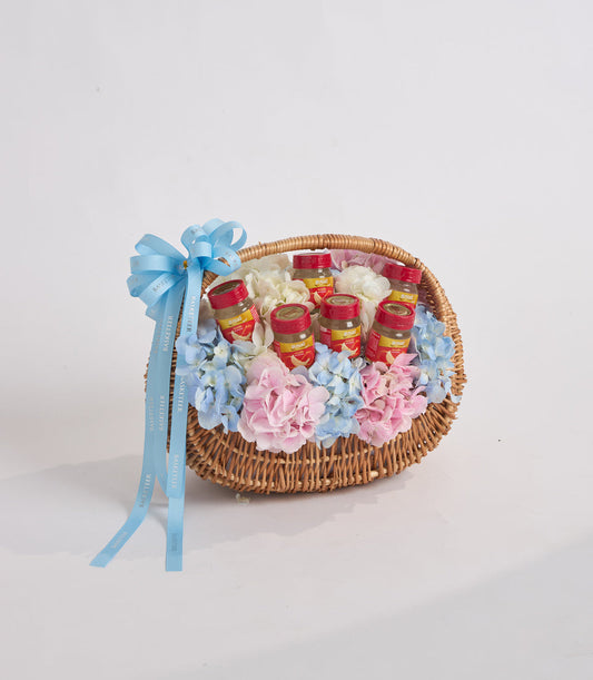 Wicker basket with red and gold jars, pink and blue flowers, blue ribbon bow on white background.