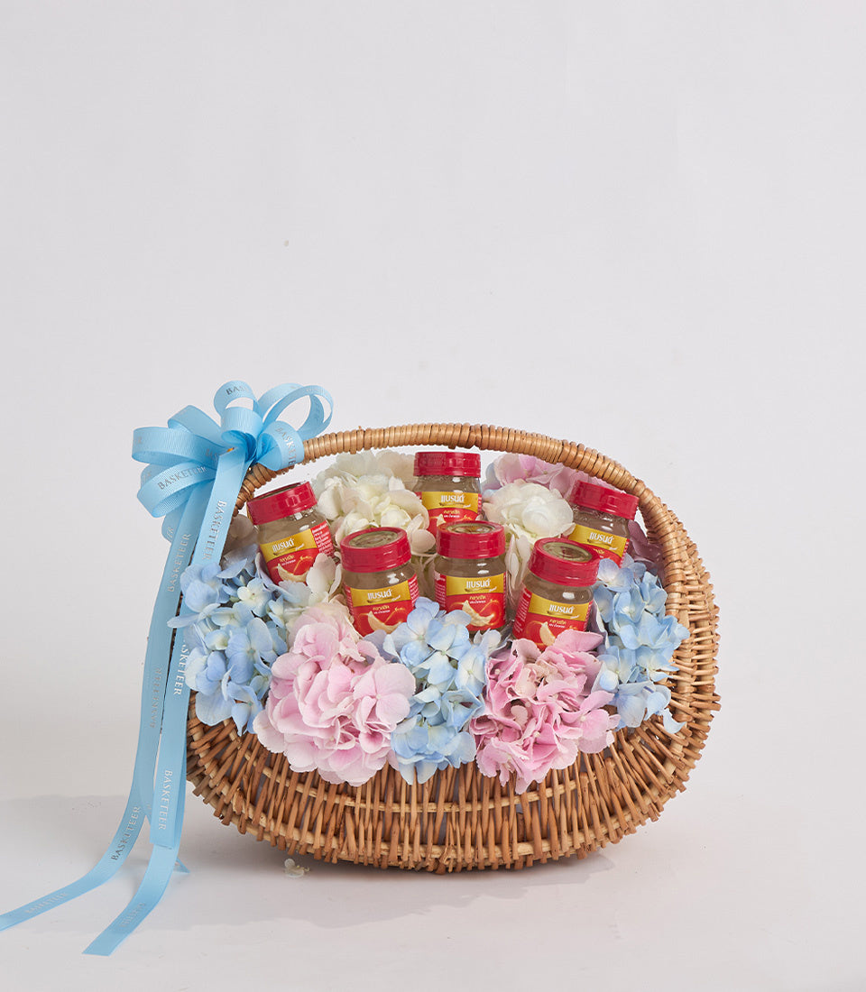 Wicker basket with blue ribbon bow containing pink and blue flowers and red condiment jars on white background.