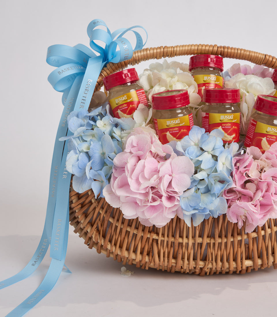 Wicker basket filled with pink and blue flowers, red-capped jars with yellow labels, and a blue ribbon bow on the handle.