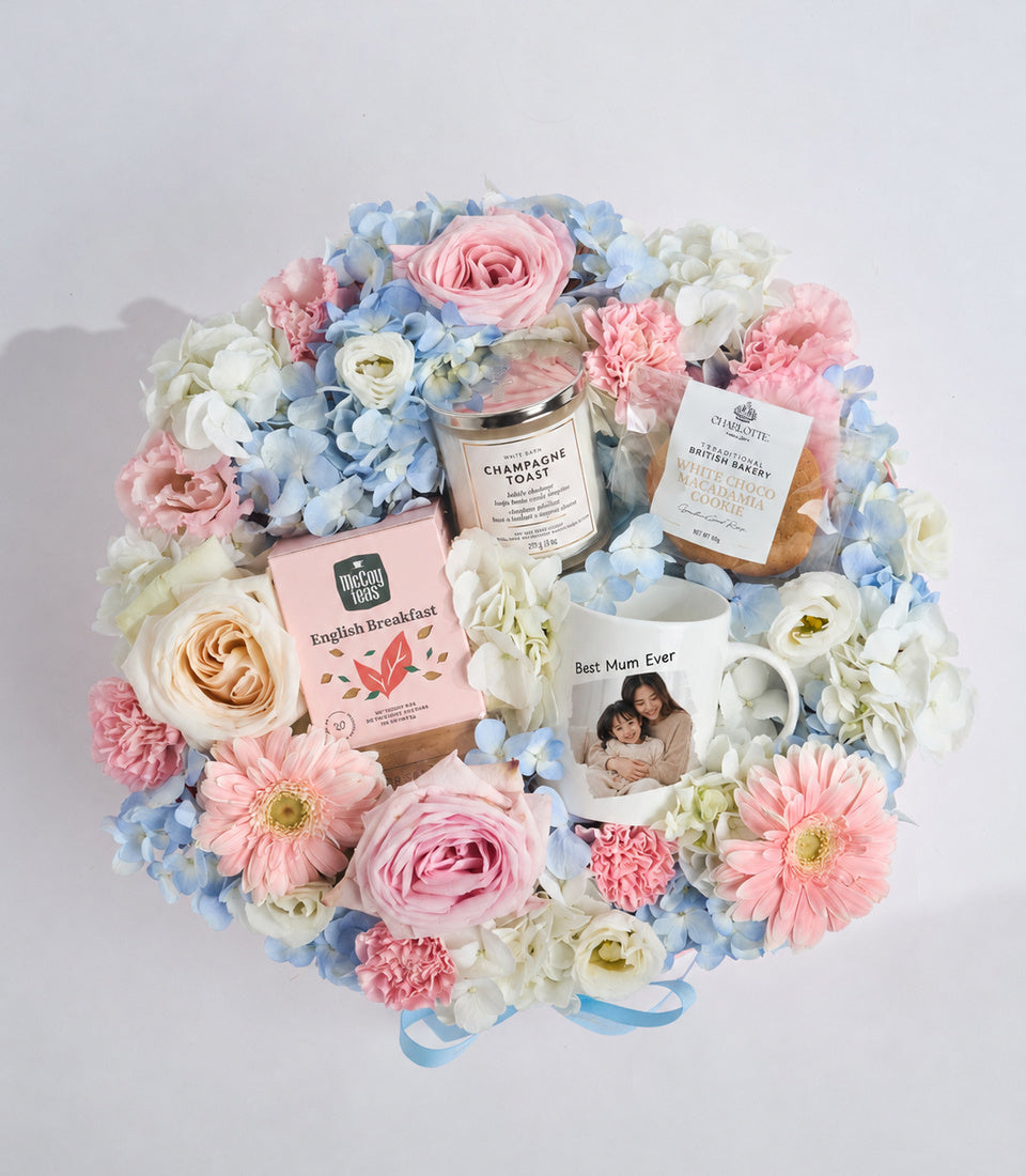 Luxury gift hamper with pink roses, white hydrangeas, and blue flowers surrounding gourmet items including tea, candle, biscuits, and personalized "Best Mum Ever" mug.