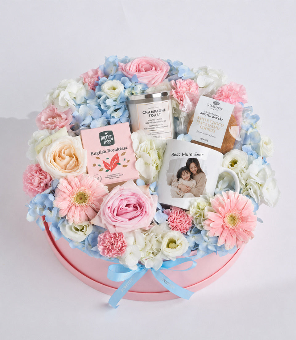 Luxe floral gift box with pink roses, white ranunculus, blue hydrangeas, pink gerberas in pink hatbox with tea, candle, cookies and personalized photo card.