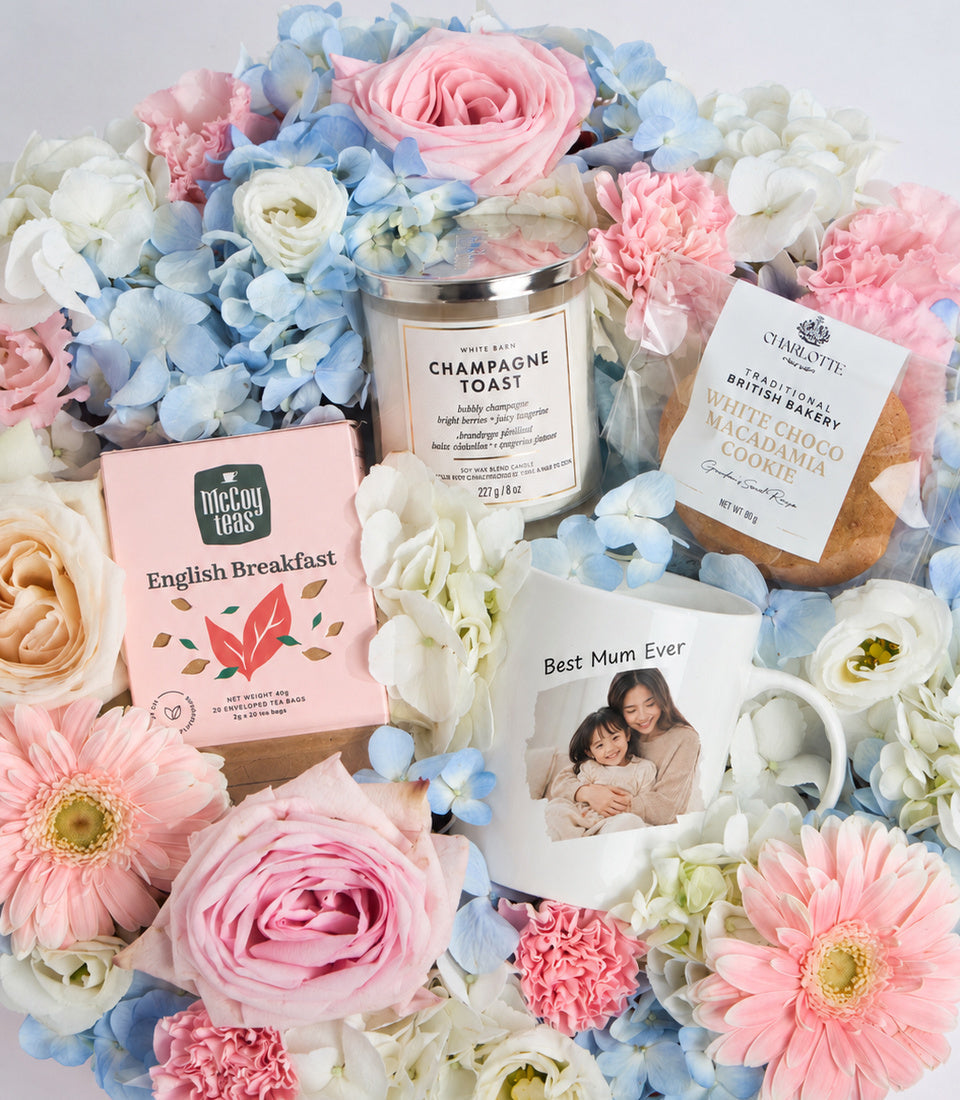 # Alt Text:

Mother's Day gift set with pink and white flowers, English Breakfast tea, Champagne Toast candle, White Choco Macadamia cookie, and personalized "Best Mum Ever" mug.