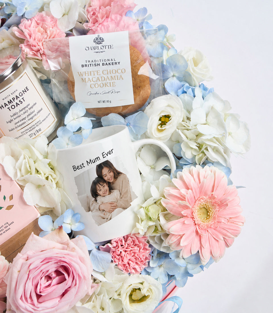 Luxury Mother's Day gift set with white mug, Charlotte bakery cookie, champagne, surrounded by pink roses, white ranunculus, and blue hydrangeas on white background.