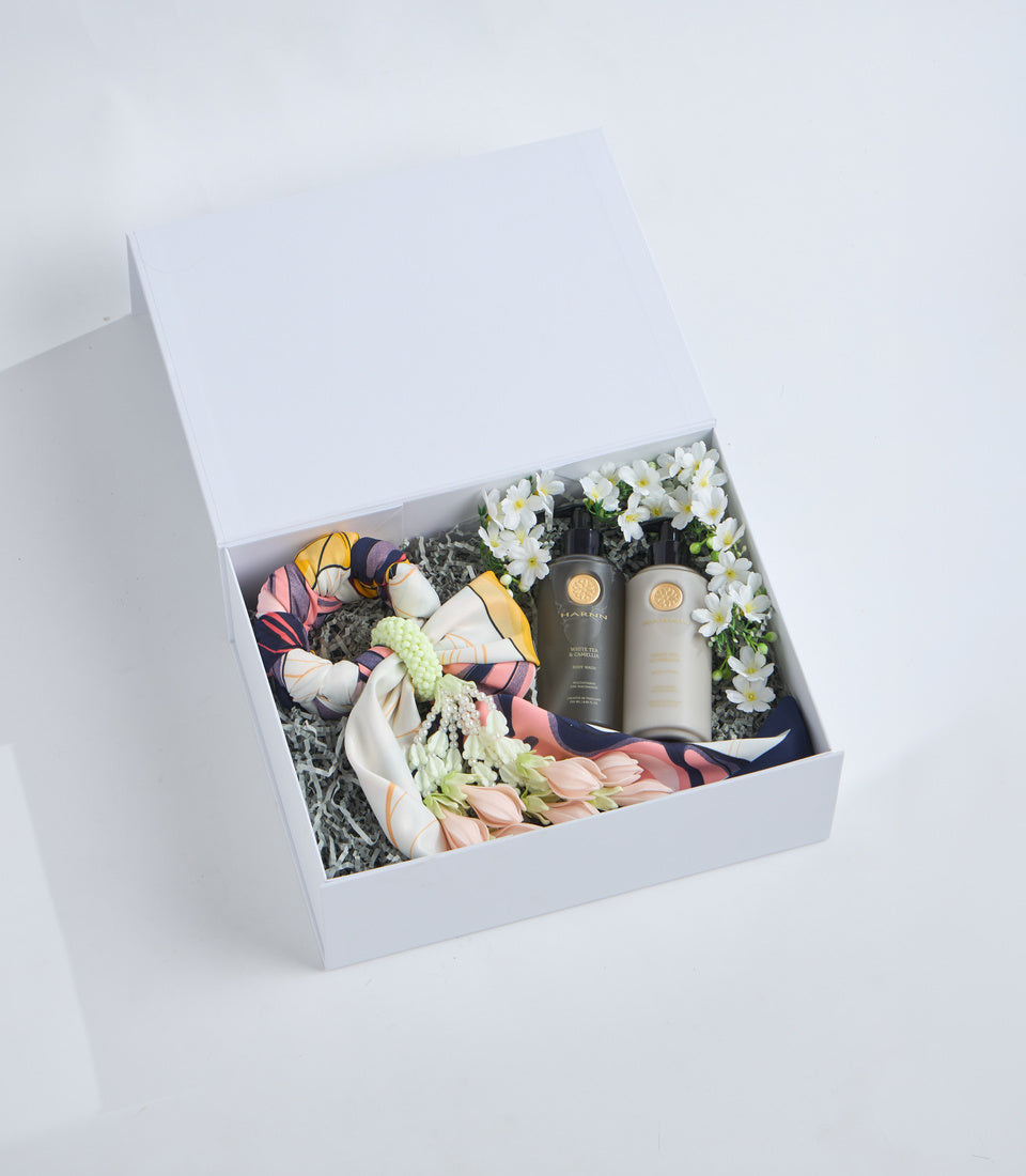 White gift box containing two beauty products, white daisies, colorful patterned silk scarf, and gray shredded paper filler on white background.