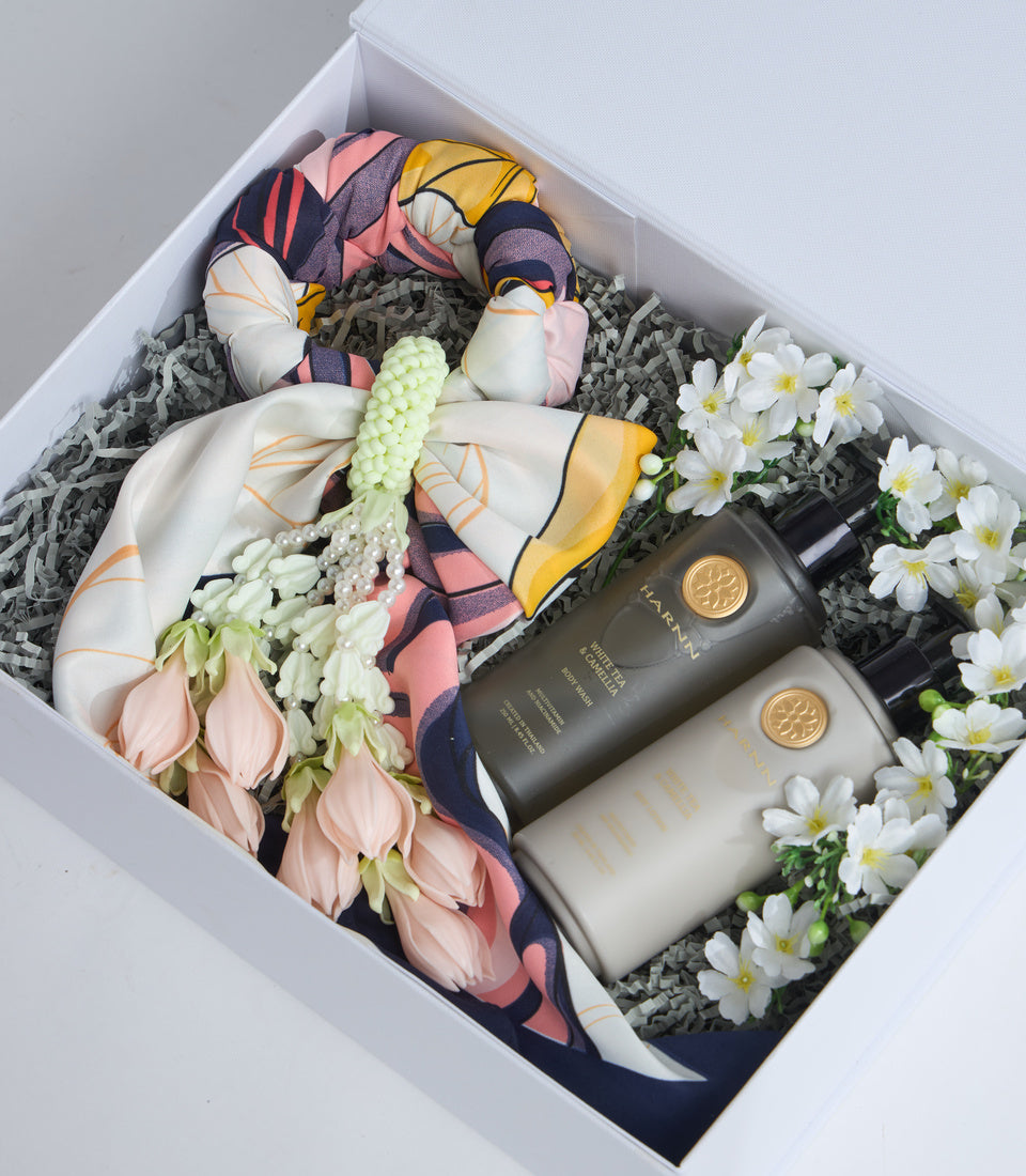Luxury gift box containing colorful silk scarves, fresh white daisies and pink flowers, beaded jewelry, and dark glass skincare bottles with gold seals.