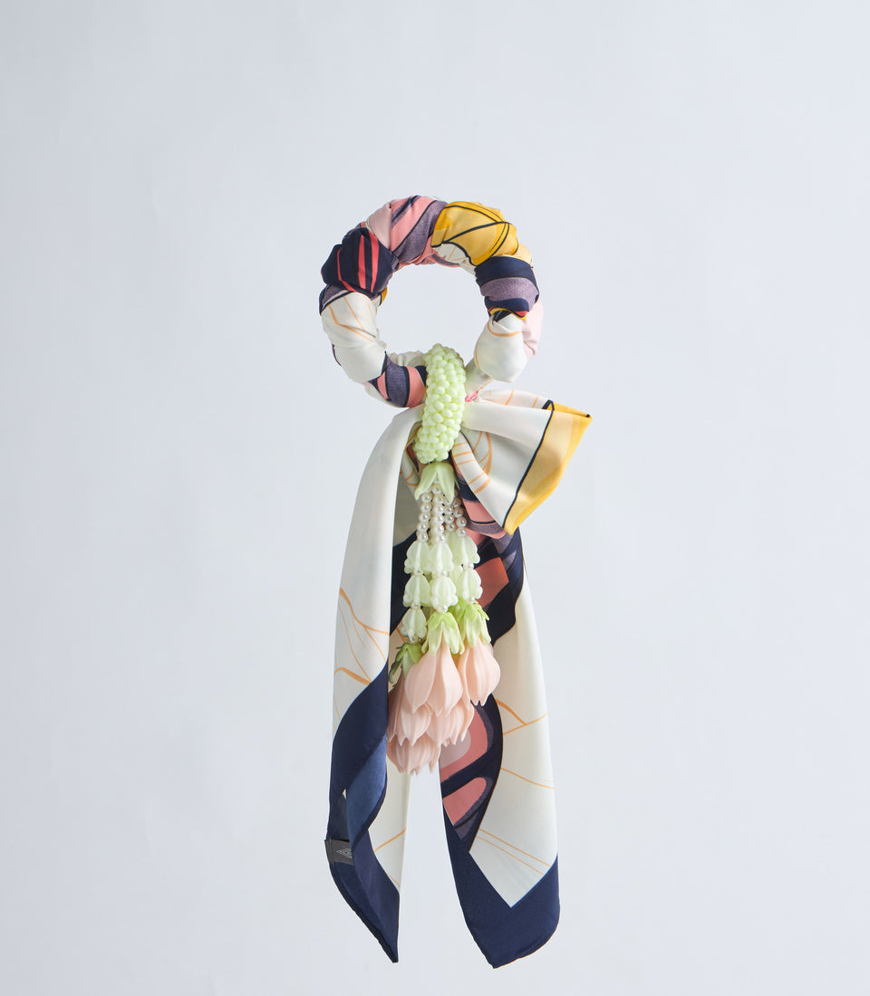 Colorful silk scarf with geometric patterns in navy, coral, yellow, and cream, adorned with fresh pink and green flowers, displayed on white background.
