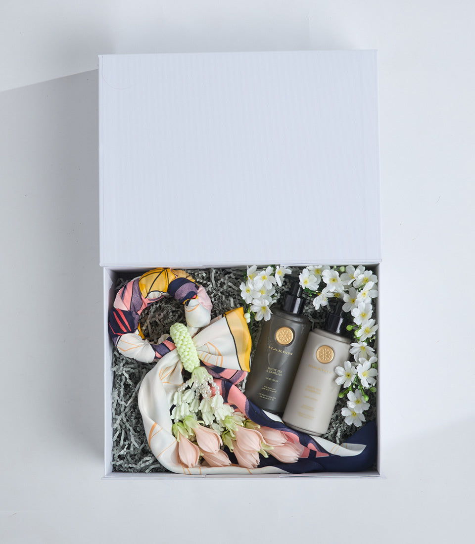 Luxury gift box containing colorful silk scarf, two dark bottles with gold seals, white daisy flowers, and green decorative filler on white background.
