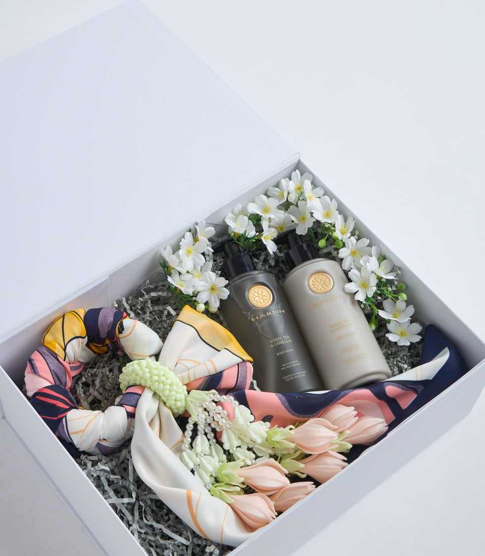 # Alt Text

Luxury skincare gift box with white daisies, pale pink tulips, colorful silk scarves, and two dark bottles with gold seals on gray shredded paper.