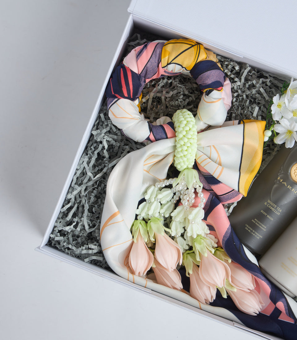 # Alt Text

Luxury gift box with colorful silk scarves, decorative ceramic pieces, pale pink tulips, white daisies, green hypericum berries, and dark cosmetic product on shredded paper filler.