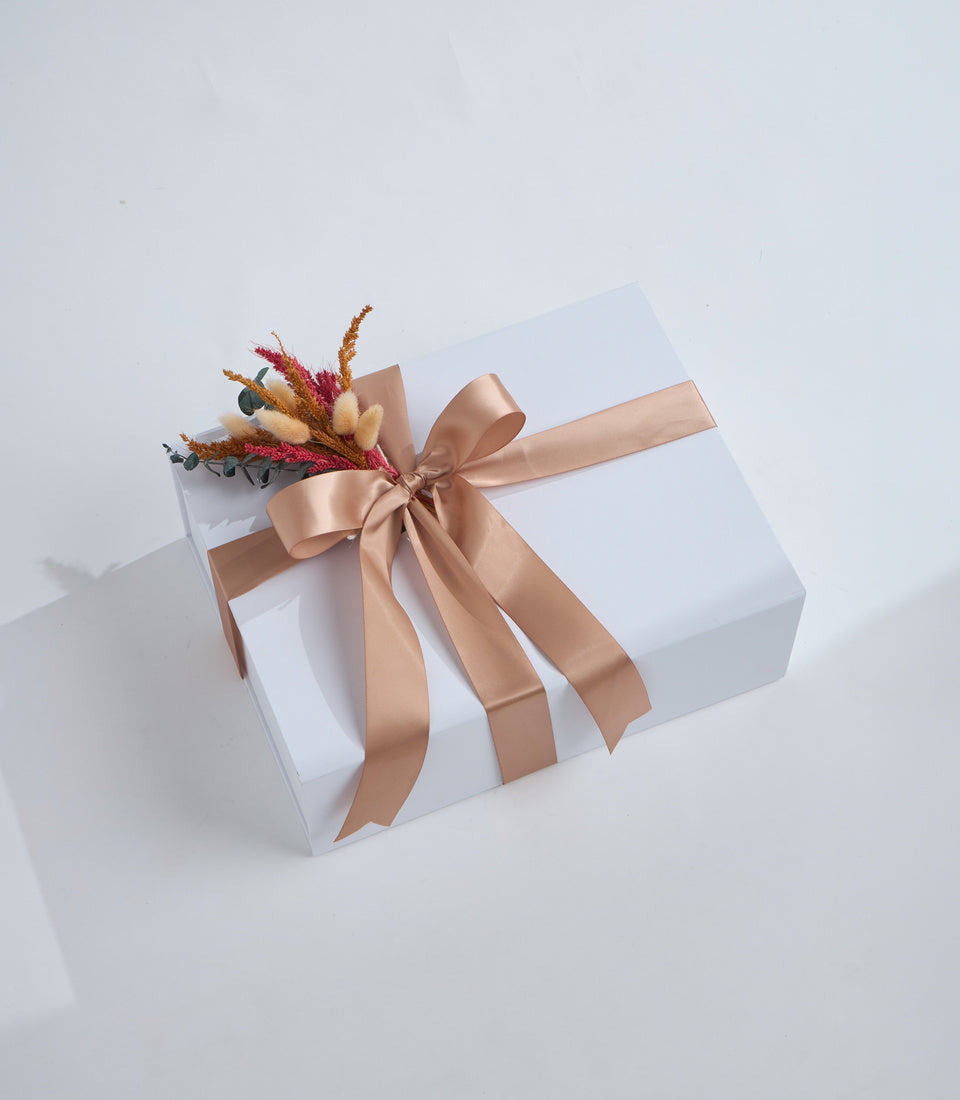 White gift box with champagne satin bow and dried flower arrangement featuring red, orange, and cream-colored blooms on light background.