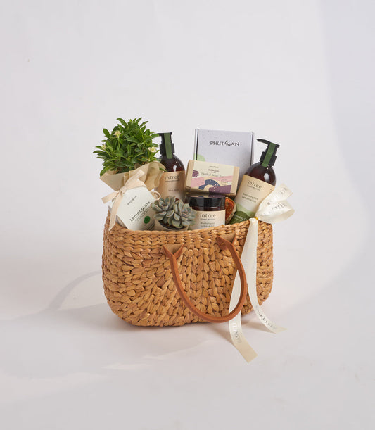 Woven straw basket filled with skincare products, green potted plants, succulents, and branded boxes with tan ribbon handles on white background.
