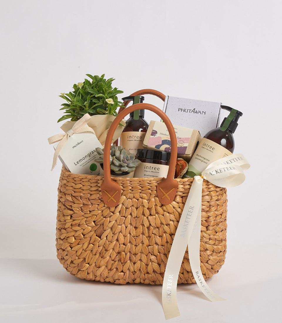 Woven straw basket with brown leather handles filled with green plants, skincare products, and wellness items on white background.