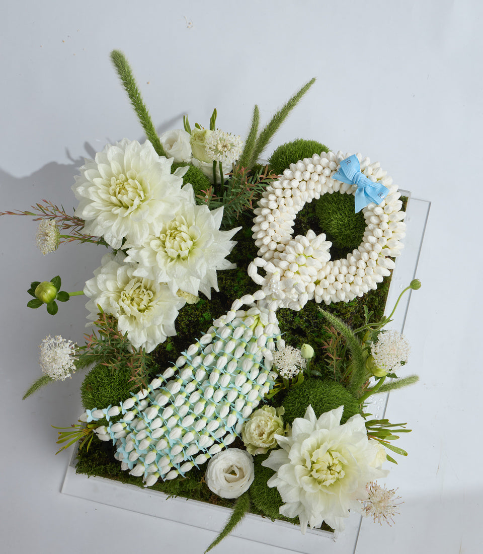 Elegant floral arrangement featuring white dahlias, pearl spray flowers, moss spheres, ornamental grasses, and a blue ribbon bow on a white base.