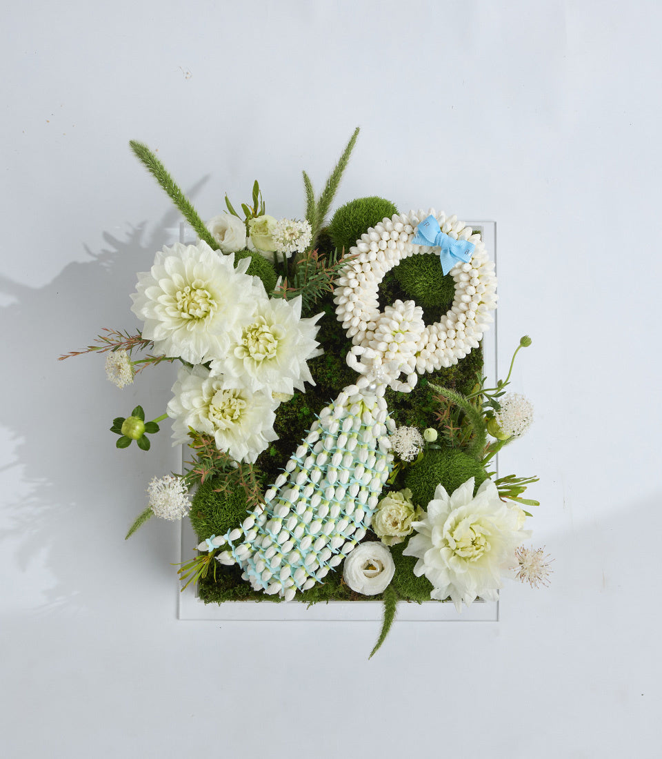 Elegant floral arrangement featuring white dahlias, baby's breath, green moss, ornamental grasses, and a light blue ribbon bow on a white square base.