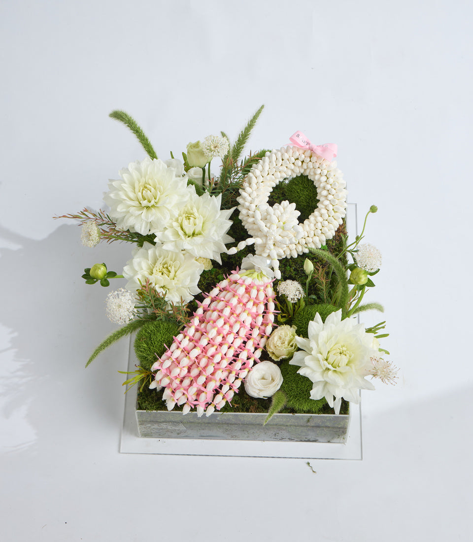 Elegant floral arrangement in rustic wooden box featuring white dahlias, pink sola flowers, green moss, and ornamental grasses on white background.