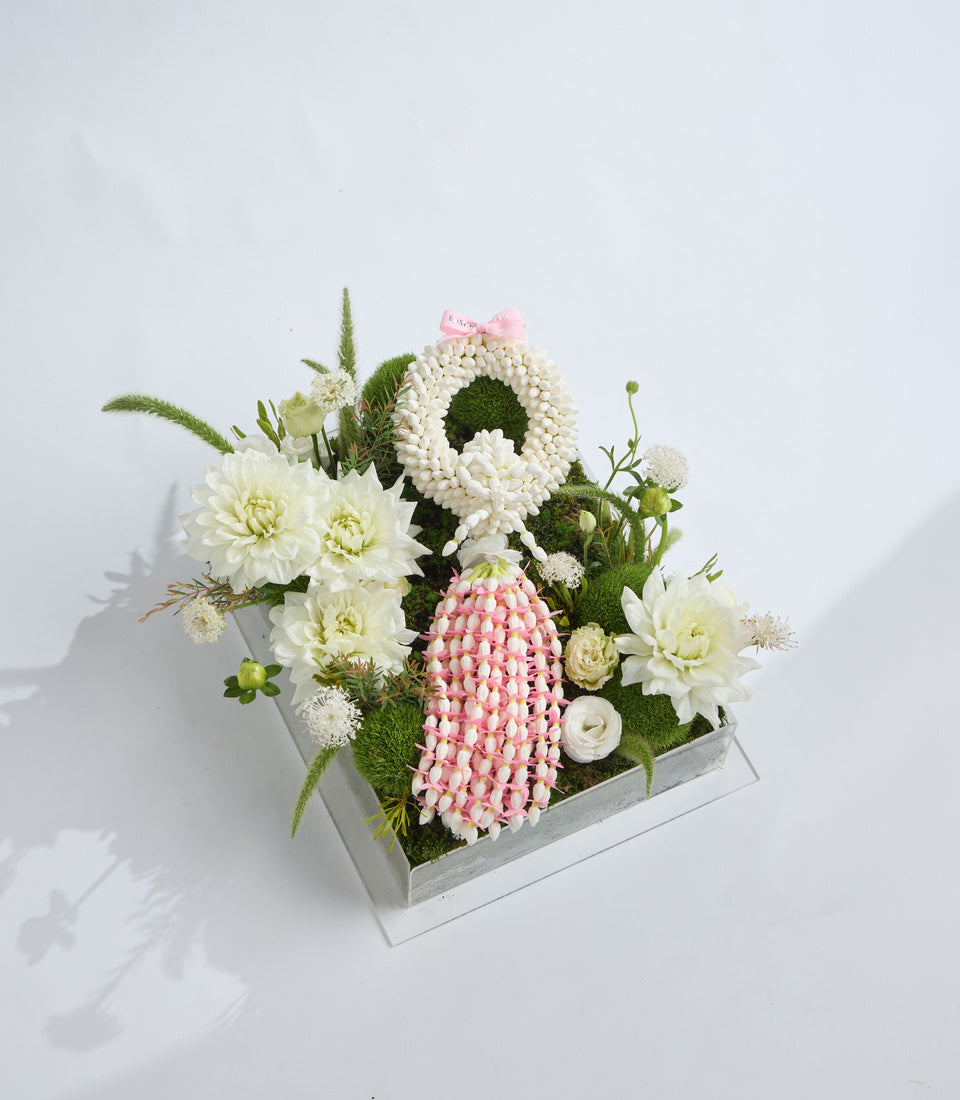 Elegant floral arrangement in white box featuring pink protea, white dahlias, cream roses, white gerbera wreath, green foliage and decorative moss on white background.