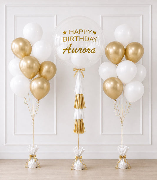 Elegant birthday balloon display featuring gold and white latex balloons with a large transparent bubble balloon, gold tassel garland, and weighted balloon bases against a white paneled wall.