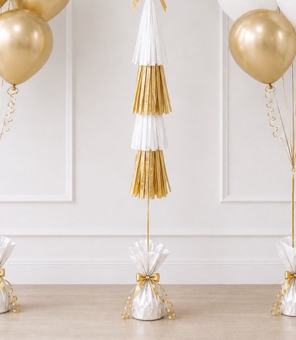 Gold and white striped tassel garland decoration with metallic balloons and gift bag accents on white wall backdrop.