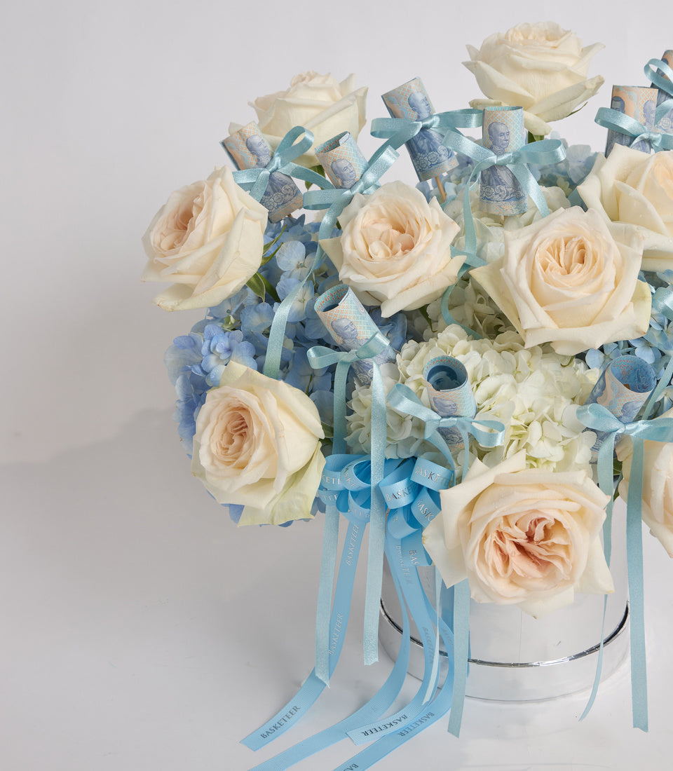 Elegant floral arrangement with cream roses, blue hydrangeas, and eucalyptus in white cylindrical vase with blue ribbon bows.