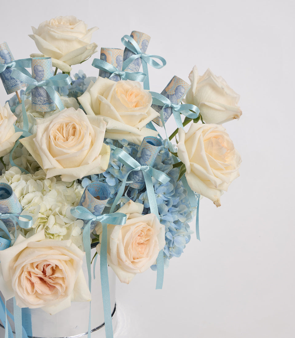 Elegant floral arrangement with cream roses, white hydrangeas, and blue decorative ribbons in a clear glass vase against a white background.