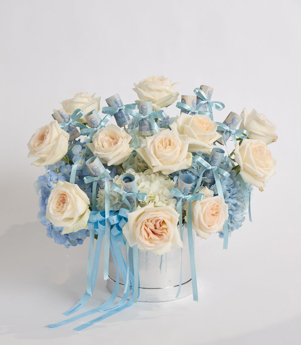 Elegant floral arrangement with cream roses, blue hydrangeas, and dusty miller in white cylindrical vase with blue ribbon bows.