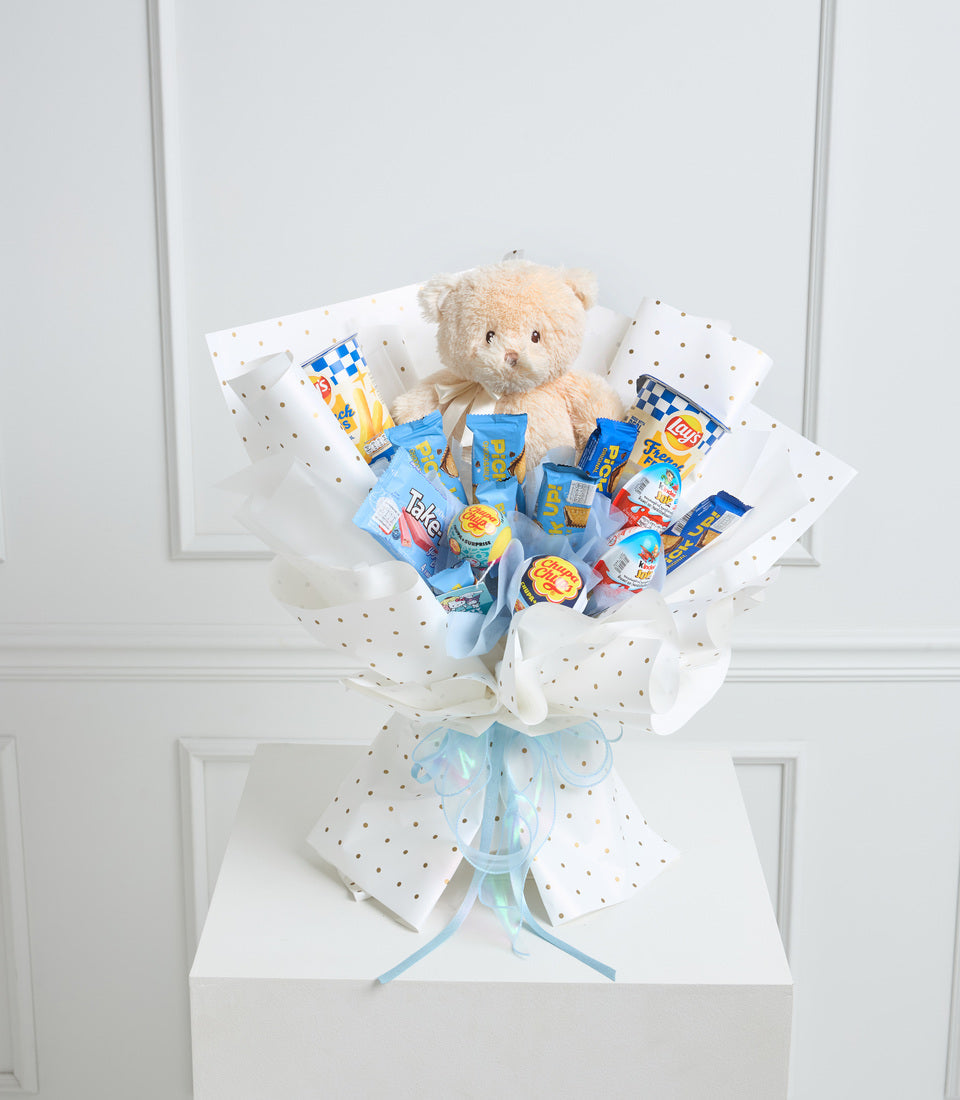 Cream teddy bear surrounded by blue snack packages in white polka-dot wrapping paper with blue ribbon bow on white pedestal.