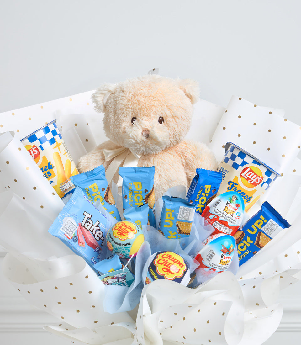 Cream teddy bear with blue ribbon surrounded by snack packages including chips and candies, arranged in white polka dot tissue paper.