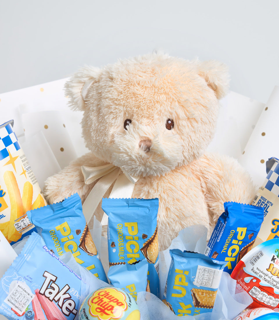 Cream teddy bear with beige ribbon bow surrounded by blue snack packets and colorful packaged treats on white background.