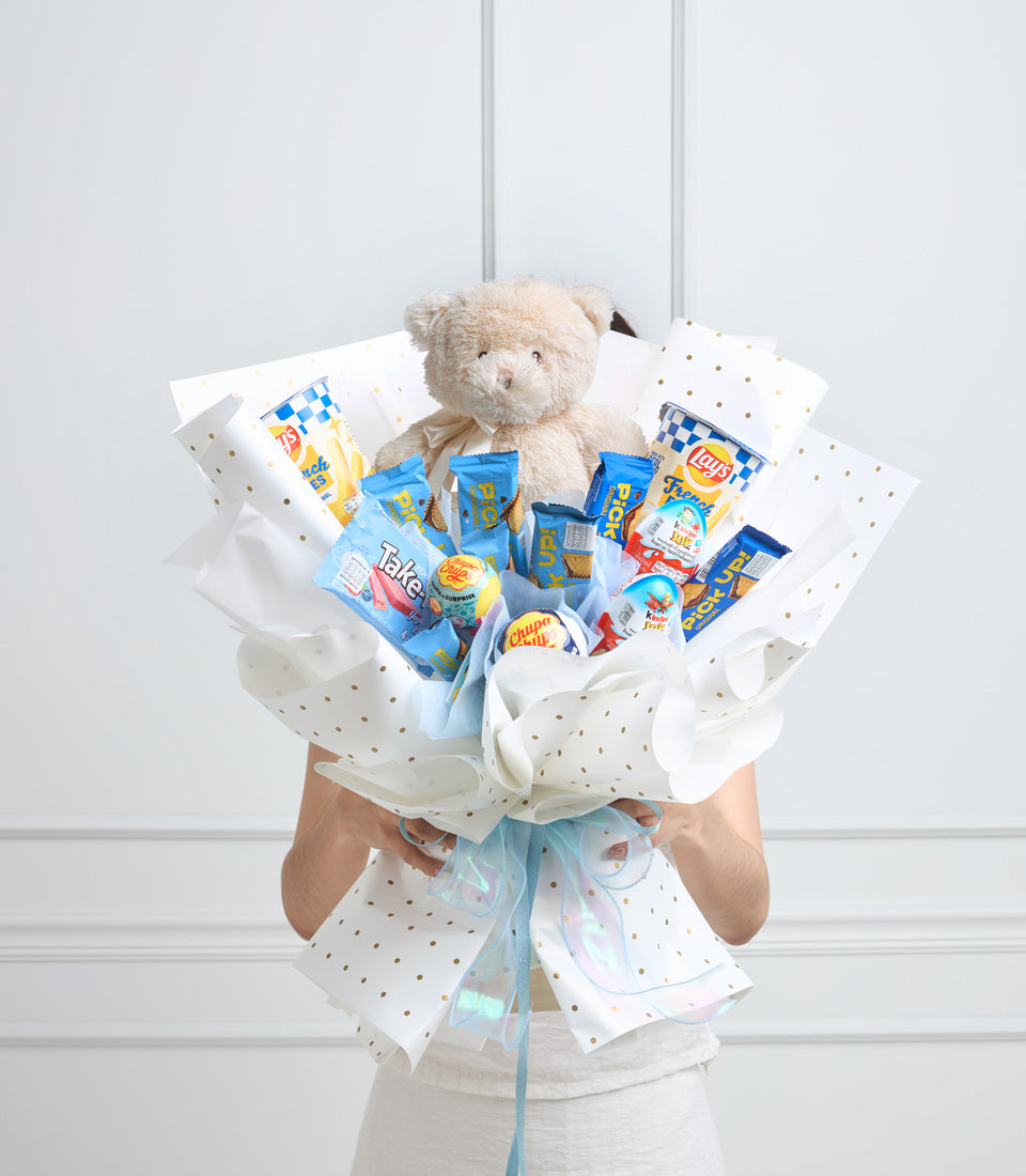 Cream teddy bear with blue bow surrounded by snack packages in white polka-dot wrapping paper with blue ribbon, held by hands against white background.