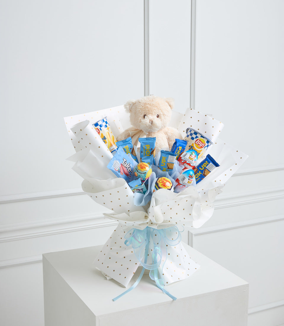 Cream teddy bear surrounded by blue snack packages in white polka-dot wrapper with light blue ribbon bow, displayed on white pedestal.