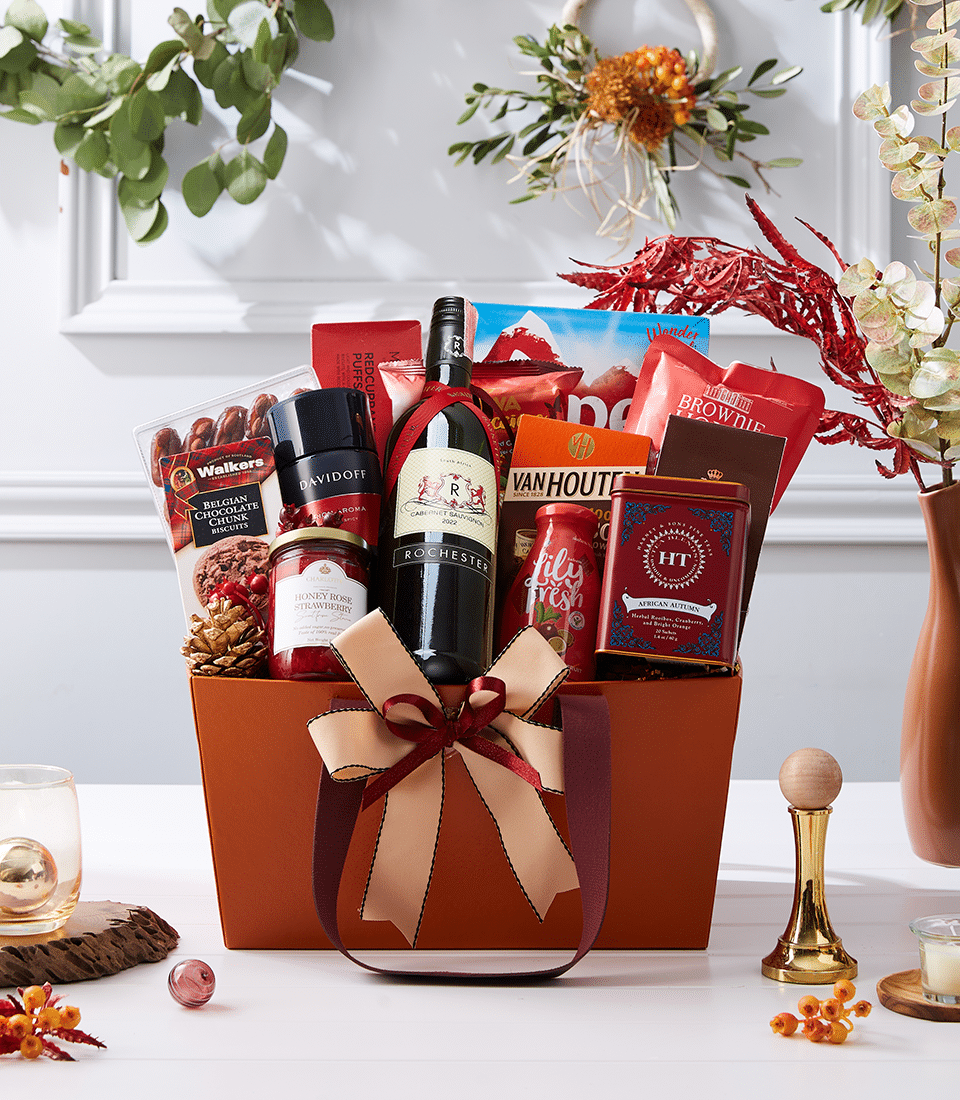 Premium red wine gift hamper with gourmet treats, strawberry jam, and artisan snacks in tan woven basket with burgundy bow