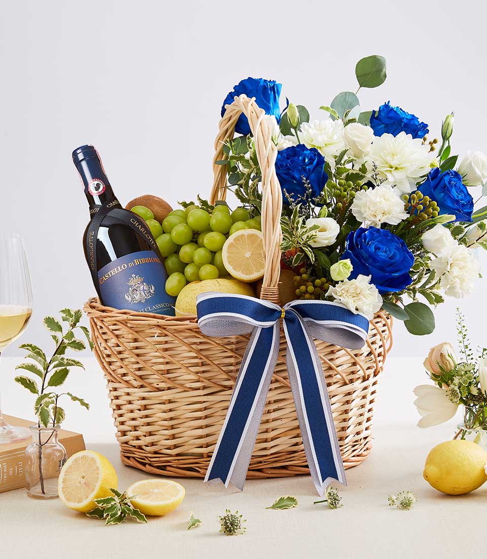 Premium red wine gift basket with blue roses, white carnations, fresh fruit, and navy ribbon bow